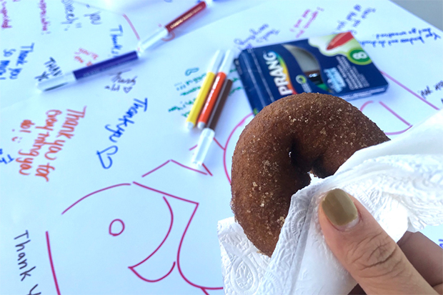 Closeup of a hand holding a cider donut in front of a poster board with colorful thank yous written over it.